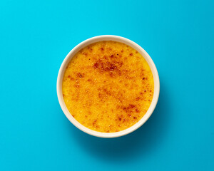 Wall Mural - Creme brulee in ramekin isolated on bright blue color background. Close up french dessert. Burned cream. Spanish crema catalana. Top view