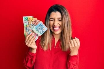 Poster - Beautiful brunette woman holding australian dollars very happy and excited doing winner gesture with arms raised, smiling and screaming for success. celebration concept.