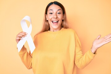 Sticker - Young brunette woman holding cancer awareness white ribbon celebrating victory with happy smile and winner expression with raised hands