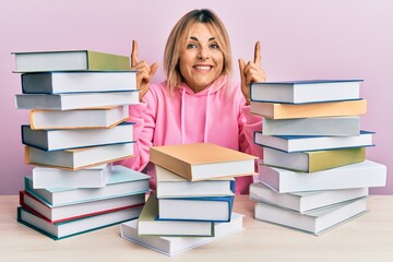 Young caucasian woman sitting on the table with books smiling amazed and surprised and pointing up with fingers and raised arms.