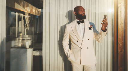 An outdoor portrait of a dapper cheerful mature bald bearded black guy in an elegant white suit, having a video call or taking a selfie via his smartphone near the glass wall of a restaurant