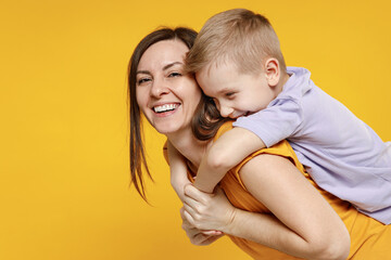 Happy young woman have fun with cute child baby boy 5-6-7 years old in violet t-shirt stand behind hug kiss Mommy little kid son together isolated on yellow background studio Mother's Day love family