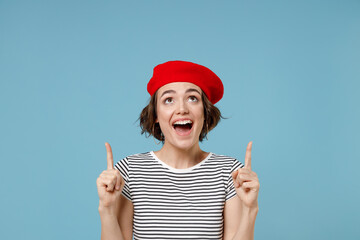 Wall Mural - Young smiling european woman 20s with short hairdo wear french beret red hat striped t-shirt point index finger overhead on workspace area mock up isolated on pastel blue background studio portrait