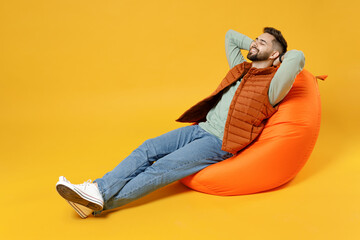 Wall Mural - Full length young smiling relaxed fun caucasian man 20s wear orange vest mint sweatshirt sitting in beanbag bag chair hold hands behind neck head resting isolated on yellow background studio portrait.
