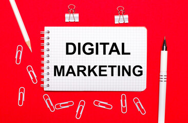 Fototapete - On a red background, a white pen, white paper clips, a white pencil and a notebook with the text DIGITAL MARKETING. View from above
