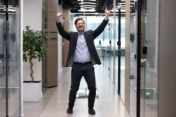 Overjoyed young Caucasian male employee dance in office hallway celebrate work success or job achievement. Happy man worker have fun or party at workplace, excited with promotion or victory.