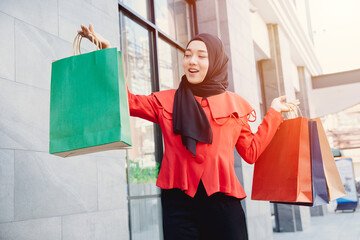 Arab Muslim young woman in veil hijab clothes holding shopping bags and walking on the city street. Shopping time. Modern skyscrapers in the background.