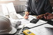 © amnaj - Close-up of a man architect holding a pen brainstorming ideas on a blueprint design using a calculator and laptop at the office.