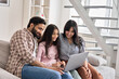 © insta_photos - Happy indian family couple with child daughter using laptop computer at home. Smiling parents and teen kid bonding watching streaming online tv or doing ecommerce shopping together sitting on sofa.