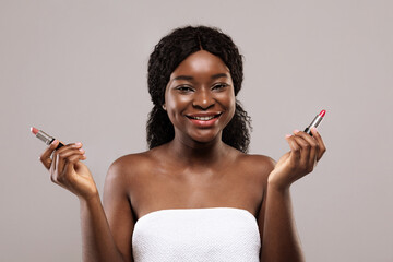 Smiling young black woman wrapped in towel holding two lipsticks in hands