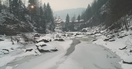 Wall Mural - Mountain river in ice and snow at sun pine forest aerial. Winter nature landscape. Bridge with tourists over waterfall. Icy water flowing over stones at spruce trees. People at resort. Alpine stream