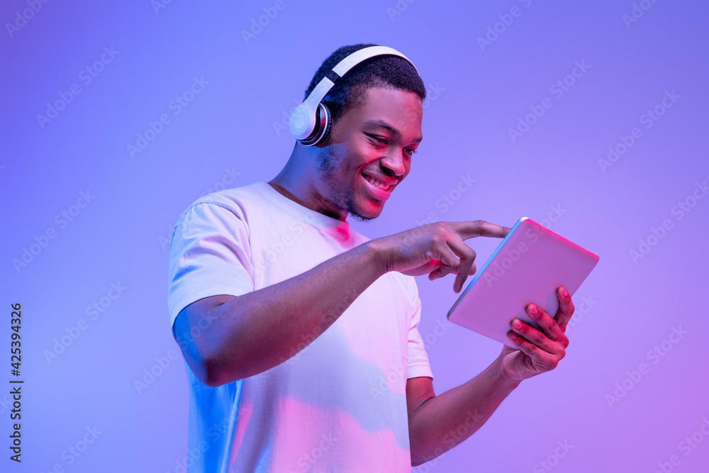 Interesting App. Excited African American Guy In Wireless Headphones Using Digital Tablet - obrazy, fototapety, plakaty 