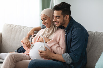 Wall Mural - Pregnancy Music. Happy Pregnant Muslim Couple Placing Wireless Headphones On Woman's Belly