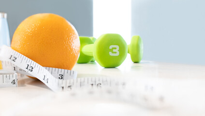 3 kilograms of green dumbbells with orange and measurement tape swirl around at blurred background, depth of field. Weight loss and wellness by personal bodybuilding and fresh fruit nutrition concept.