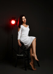 Sensual and wise adult woman in white dress sits barefooted on chair in neon spotlight glow over dark background. Fashion, stylish casual look for lady concept
