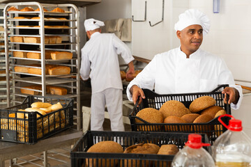 Wall Mural - Positive man baker carrying tray with fresh baked bread at bakery kitchen