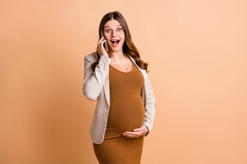 Poster - Photo of pretty excited woman expecting child wear jacket arm belly talking modern device isolated beige color background