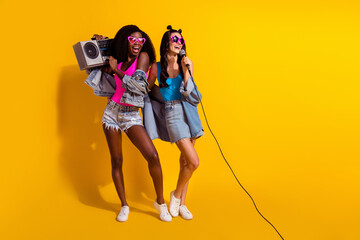 Photo of two cute ladies wear jeans outfit dark eyewear listening boom box singing looking empty space isolated yellow color background