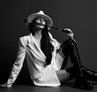 Black and white. Gorgeous woman vamp mistress in white hat, white jacket and black high leather boots sits holding horse whip, riding crop in mouth. Fashion, vogue, sexy stylish look for woman concept