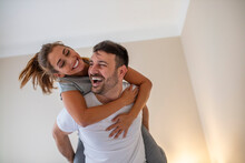 Lovely Young Couple Having Fun In The Room. Excited Young Woman Piggyback Man In Bedroom. Loving Couple Having Fun Together At Home, Laughing Spouses In New Own Apartment