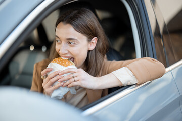 Happy woman eating a burger in the car. Bites a sandwich. Have unhealthy fast food snack. Food to go. Hungry and busy concept.