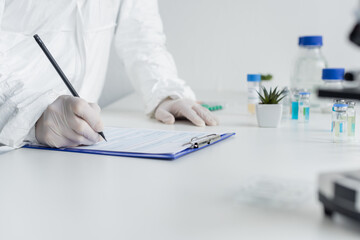 Wall Mural - Cropped view of scientist in hazmat suit and latex gloves writing on clipboard near vaccines on blurred foreground