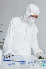 Wall Mural - Scientist in hazmat suit standing near vaccines and clipboard on blurred foreground in laboratory