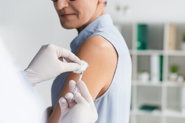 Wall Mural - Cropped view of doctor in latex gloves holding cotton and syringe near woman on blurred background