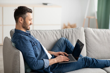 Wall Mural - Side view of bearded man reclining on couch, using laptop