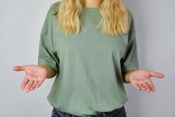 Wall Mural - Blond hair woman posing near a light wall. Beautiful young caucasian girl. Hand gestures. Emotion. Casual clothing. Studio model in work. Strong woman, future is female. Jeans and green blank t-shirt