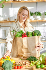 smiling caucasian woman holding shopping paper bag with fruits and vegetables stand in modern kitchen