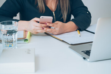 Wall Mural - Cropped photo of unrecognizable female teacher sitting at home office desk and holding mobile phone. Young professor or student having break with online class and using smartphone to send text message