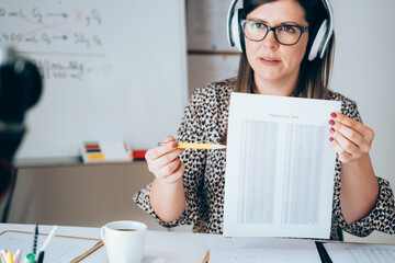 Wall Mural - Professor looking and talking direct to video camera and showing paper. Female teacher holding online class for e-learning students, recording math lesson using camera and headset for live streaming 
