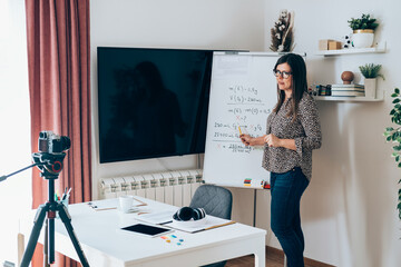 Wall Mural - Teacher recording video tutorial for e-learning class. Female professor standing and explaining the assignment pointing on the whiteboard and using camera for live streaming on the internet online