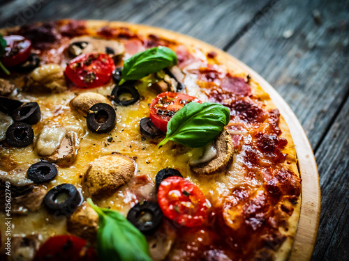 Obraz Dibond Pizza with white mushrooms, ham, tomatoes, olives, parmesan and mozzarella on wooden background