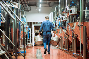 Sticker - Back view of male worker of contemporary beer production plant carrying two cisterns while moving along rows of huge steel tanks