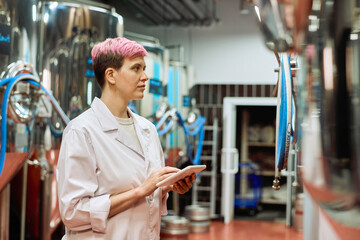 Sticker - Young female brewery expert in whitecoat entering data in tablet while looking at huge new steel equipment for beer production in plant