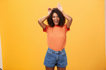 Let me be your bunny. Portrait of charming enthusiastic and charismatic happy dark-skinned female with afro hairstyle holding palms on head like animal ears making cute face over orange background