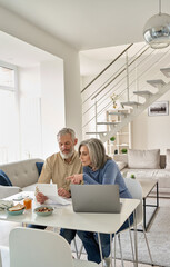 Senior mature couple check bank loan documents using laptop at home. Middle aged old family read paper bills payments, calculating pension or taxes, planning retirement finances, household expenses.