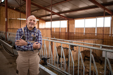 Wall Mural - Portrait of senior farm worker with arms crossed standing in farmhouse. In background domestic animals eating. Organic food and meat production.