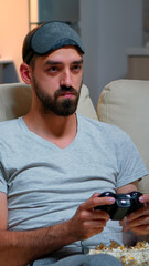 Wall Mural - Close up of man with eye sleep mask playing videogames with joystick for gaming competition. Caucasian male sitting in front on television on sofa late at night in kitchen