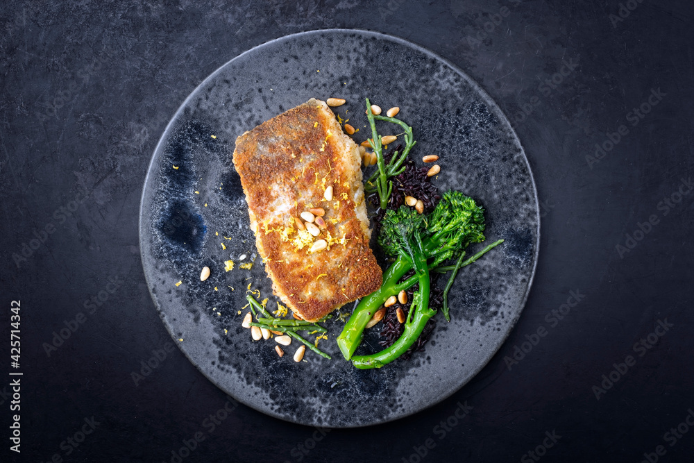 Modern style traditional pan-fried skrei cod fish filet in breadcrumbs with baby broccoli, black rice and roasted pine nuts served as top view on ceramic design plate with copy space - obrazy, fototapety, plakaty 