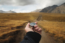 Man's Hand Holding A Magnetic Compass First-person View Against The Background Of A High-altitude Path And Mountains