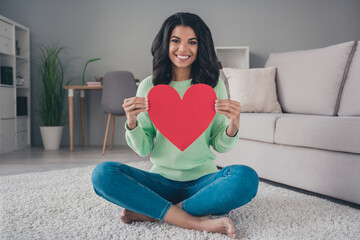 Poster - Full size photo of young cheerful black woman sit floor happy smile hold big red paper hear love valentine day home