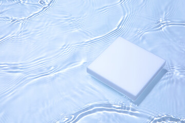 Empty white square podium on transparent clear calm blue water texture with splashes and waves in sunlight. Abstract nature background for product presentation. Cosmetic mockup, copy space.