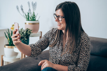 Wall Mural - Happy businesswoman sitting on sofa and using mobile phone for video call meeting. Smiling business sitting at sofa and holding smartphone for selfie or teleconferencing with colleagues from home