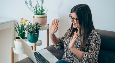 Wall Mural - Smiling businesswoman say hello to teamwork colleagues in video conference meeting while sitting on couch at home. Happy business woman with glasses looking at laptop screen and waving with hands