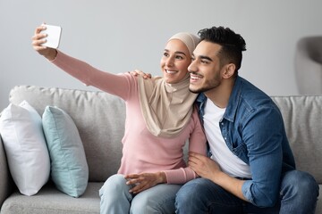 Wall Mural - Arab man and woman in hijab smiling at smartphone camera