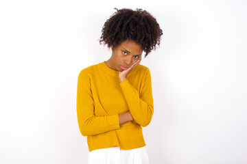 Wall Mural - Very bored young beautiful African American woman wearing yellow sweater against white wall holding hand on cheek while support it with another crossed hand, looking tired and sick.