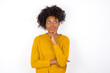 © Roquillo - Thoughtful young beautiful African American woman wearing yellow sweater against white wall holds chin and looks away pensively makes up great plan